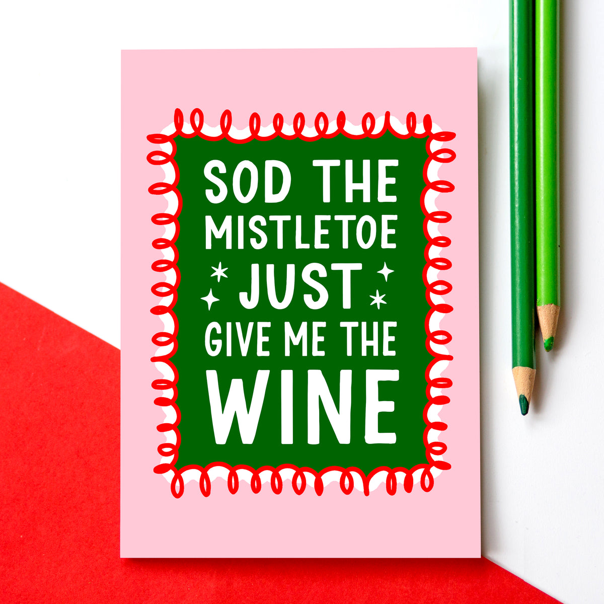 A pink christmas card with a green central panel and white text reading 'SOD THE MISTLETOE JUST GIVE ME THE WINE', styled on a white surface with a red paper corner and two green pencils.