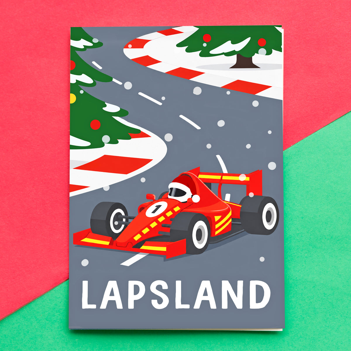 A rectangular Christmas card featuring a stylized winter race scene with a red race car on a gray road, Christmas trees, candy cane curbs, and the word "LAPSLAND" at the bottom. The item is photographed against a split red and green background.
