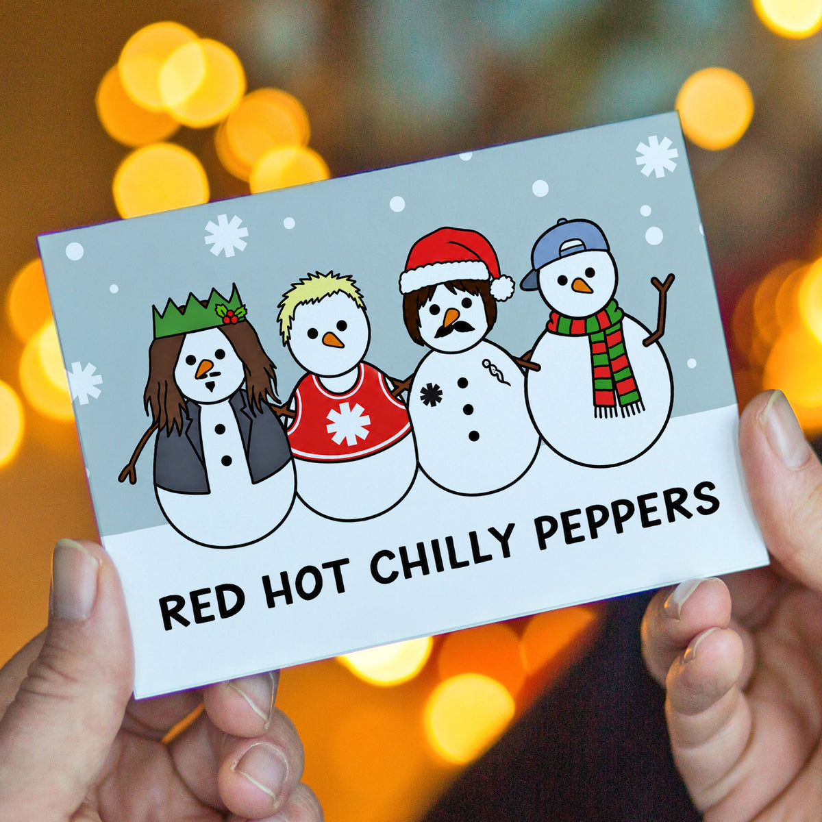 A person holds a rectangular Christmas card featuring four cartoon snowmen with winter clothing and the text 'RED HOT CHILLY PEPPERS' against a blurred background of warm yellow bokeh lights.