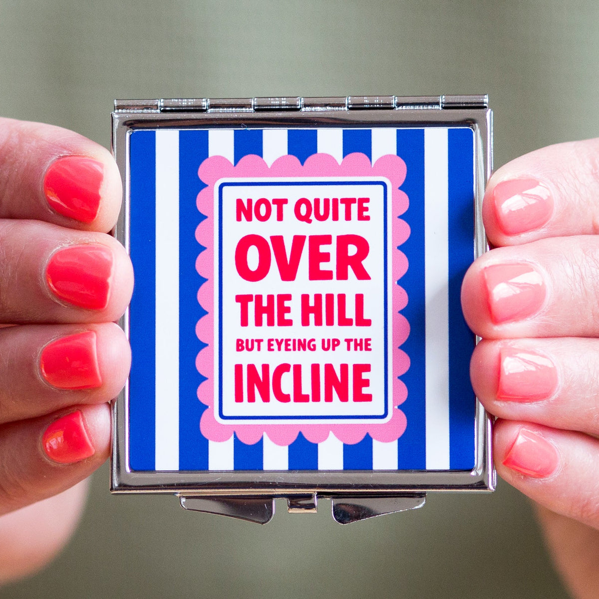 Compact mirror with blue and pink striped design held by hands with red nail polish.