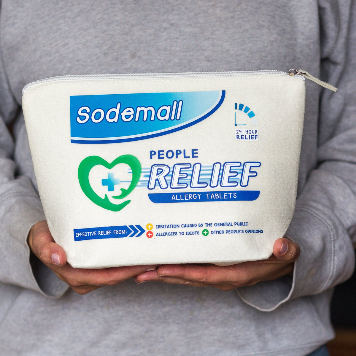 A person in a gray sweatshirt holds a white zippered pouch labeled Sodemall People Relief Allergy Tablets. The pouch features blue and green branding and text, with the person's hands and torso visible holding the product.