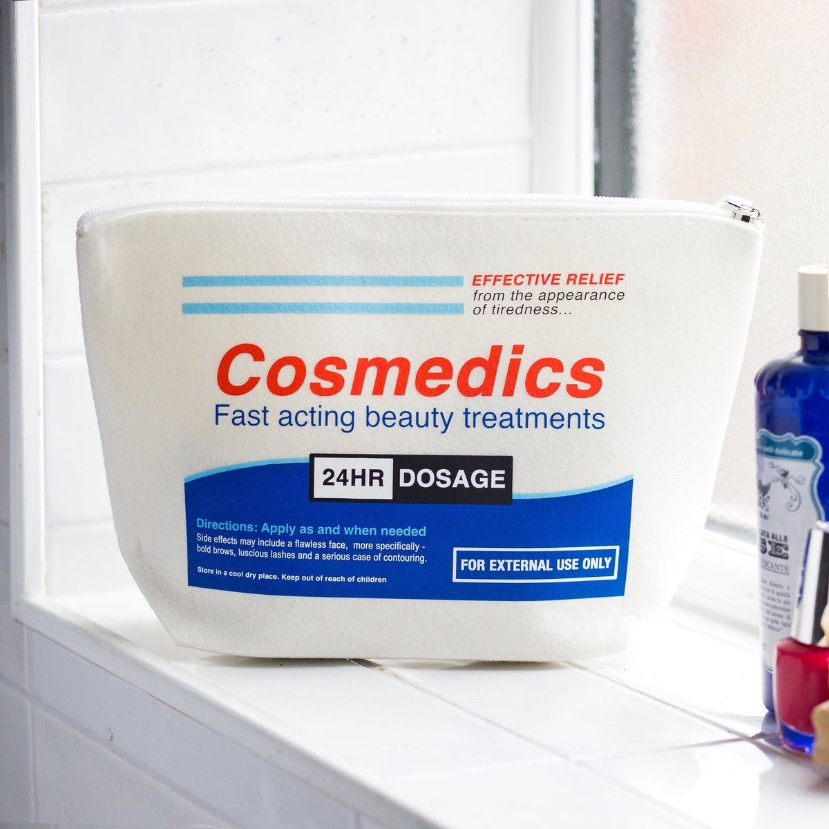 A white Cosmedics 24HR Dosage beauty treatment pouch sitting on a white tiled windowsill, with a blue bottle partially visible on the right.