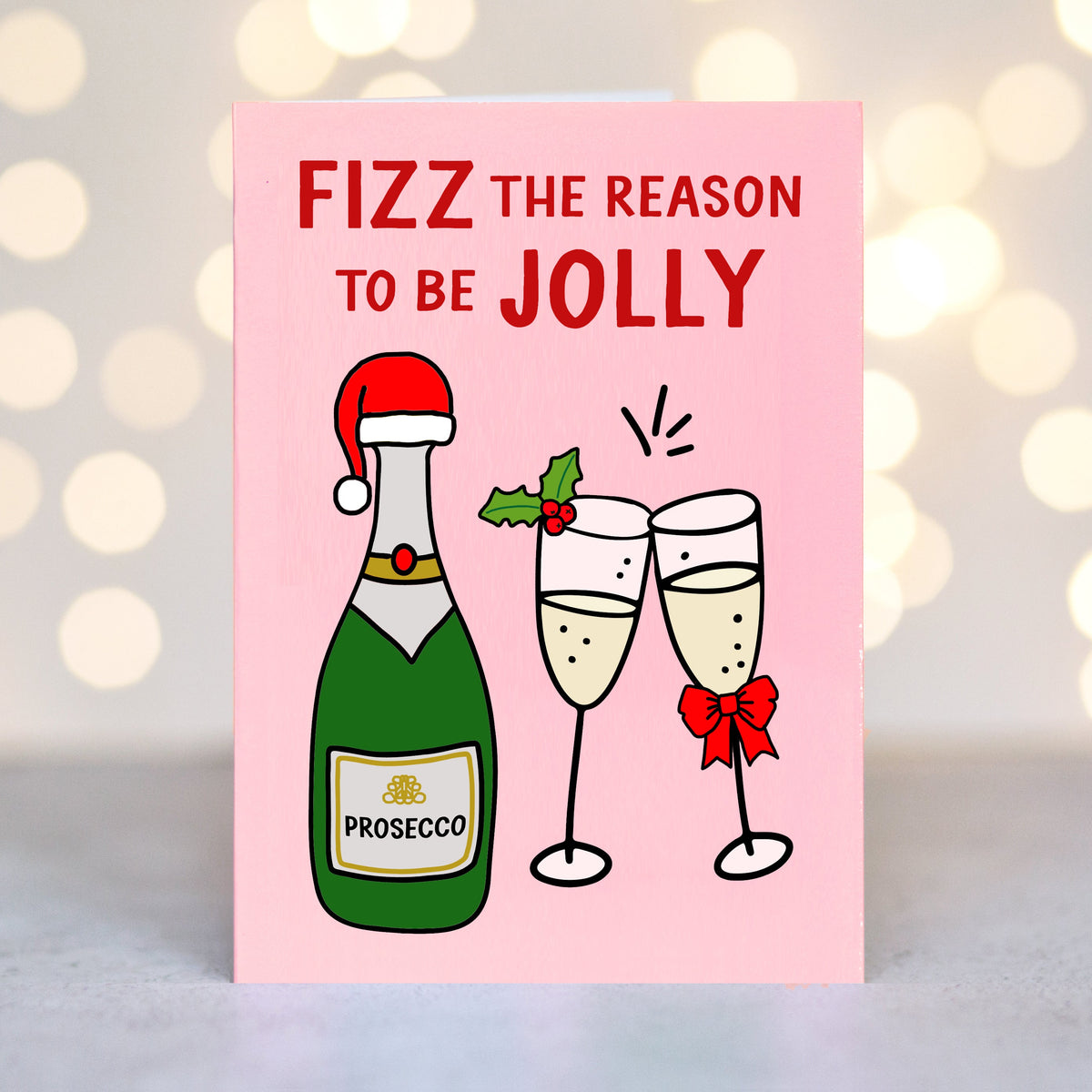 A pink Christmas card with the text 'FIZZ THE REASON TO BE JOLLY' above an illustration of a green Prosecco bottle with a Santa hat and two clinking champagne glasses, shown against a softly blurred warm bokeh background.