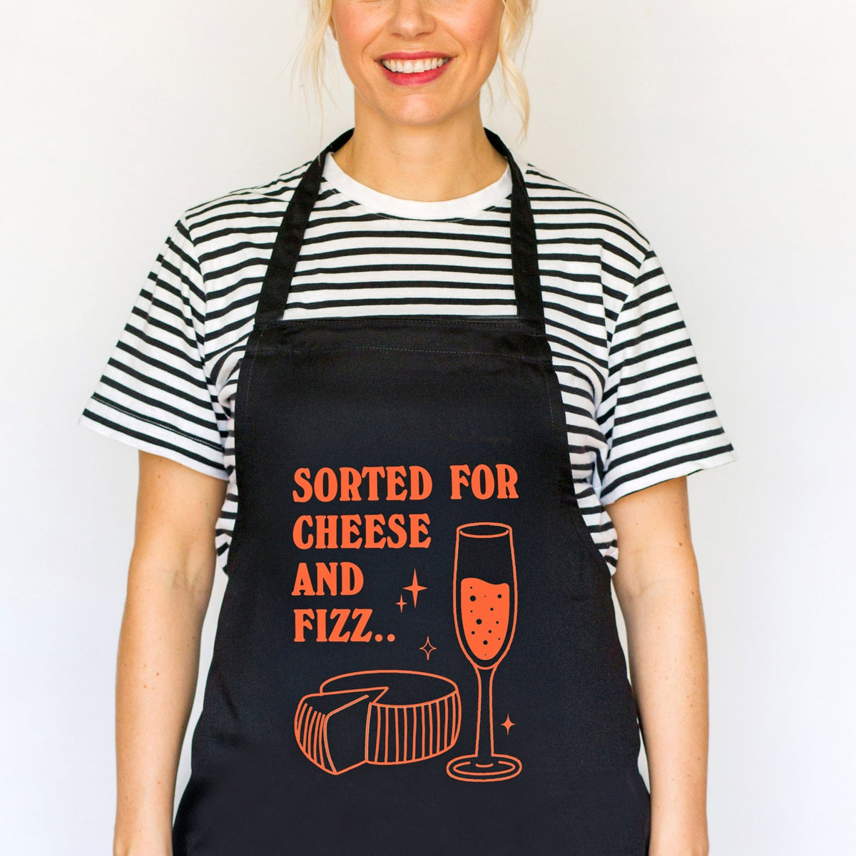 A woman wearing a black apron with orange text that reads 'SORTED FOR CHEESE AND FIZZ.