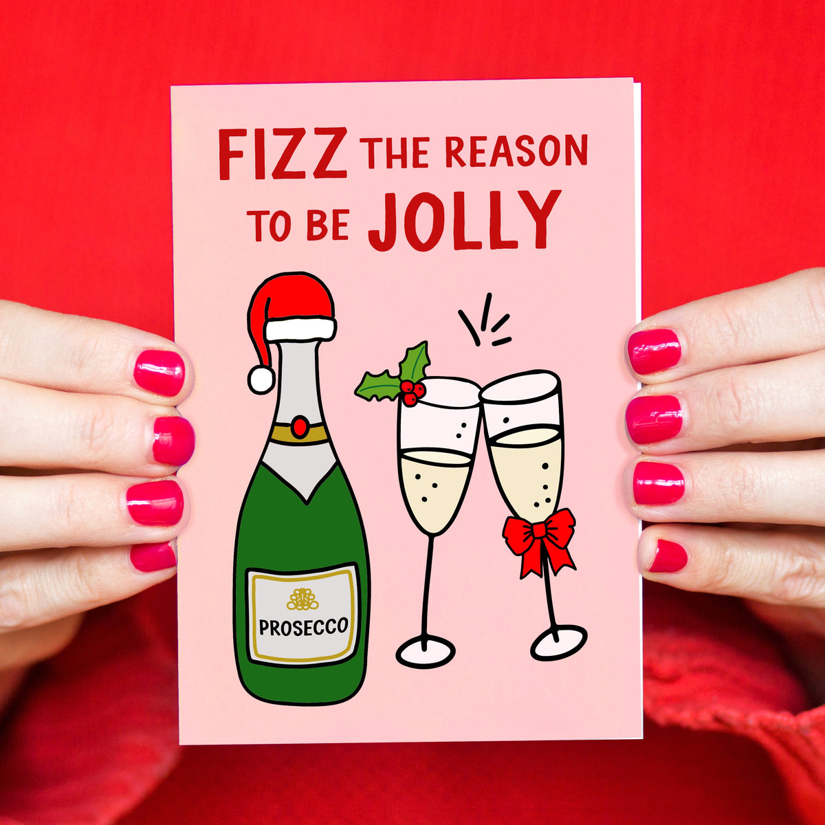 A pink Christmas card held by two hands with bright pink fingernails. The card features the text 'FIZZ THE REASON TO BE JOLLY' above an illustration of a green Prosecco bottle with a Santa hat and two clinking champagne glasses decorated with holly and a red bow.
