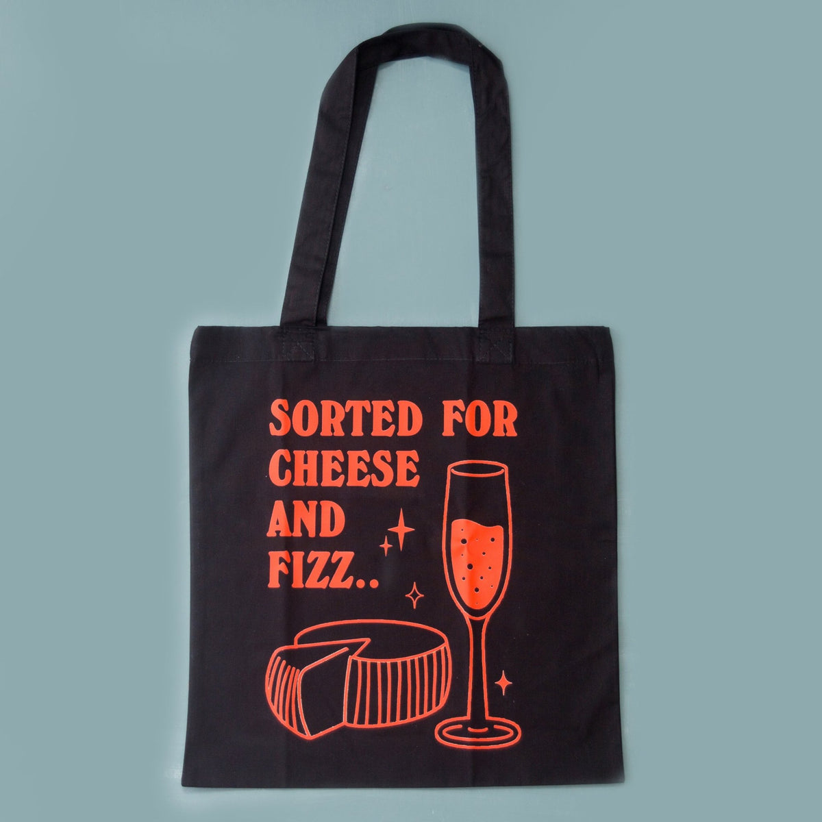 A black canvas tote bag with orange-red printed text reading 'SORTED FOR CHEESE AND FIZZ.