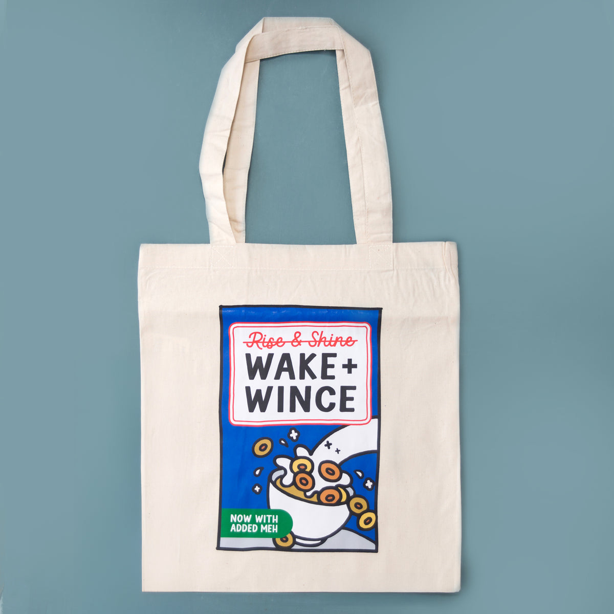 An off-white canvas tote bag with a printed parody graphic of a cereal box labeled 'Rise & Shine WAKE+ WINCE' against a solid blue background.