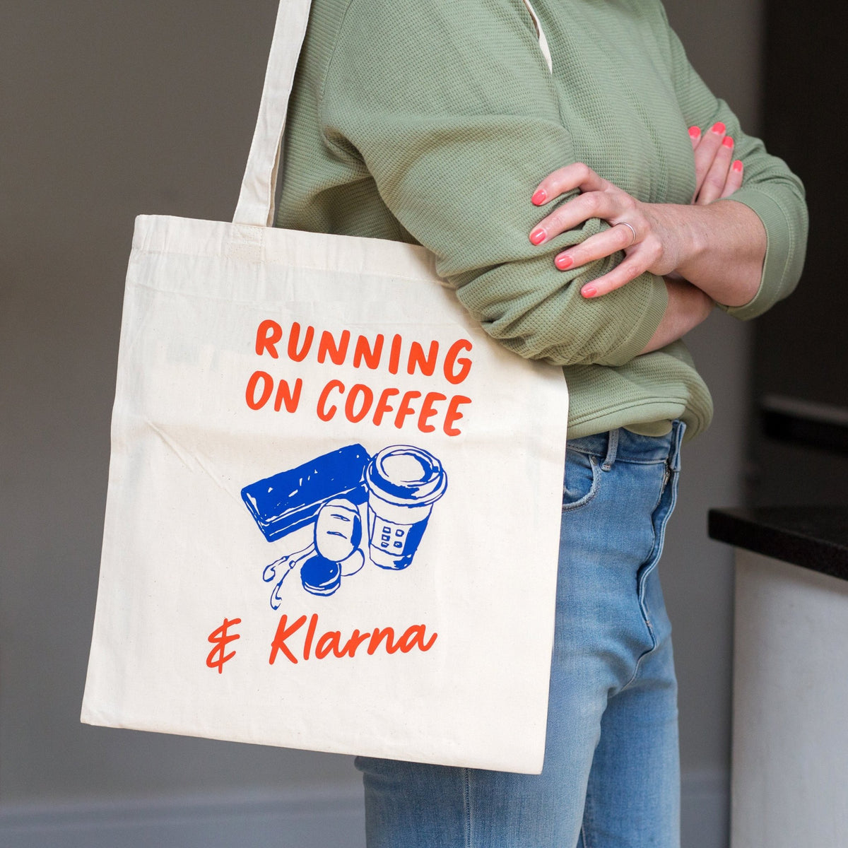A person wearing a sage green sweatshirt and blue jeans carries a cream canvas tote bag with orange and blue graphics that read 'RUNNING ON COFFEE & Klarna'.