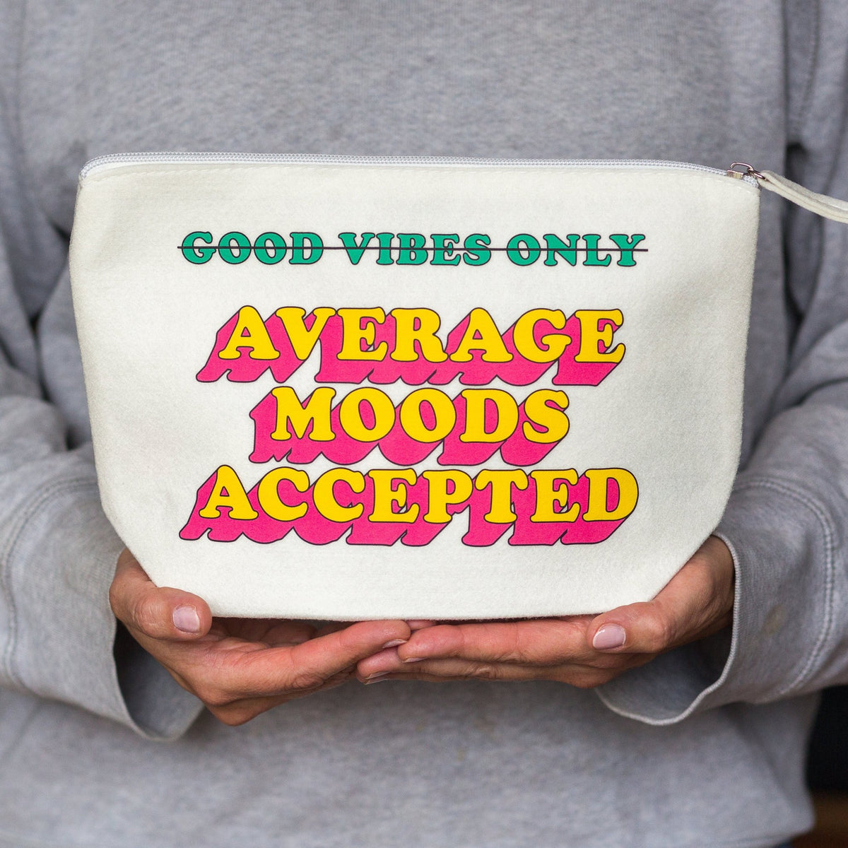 A person in a gray hoodie holds a cream-colored zippered pouch with retro-style text that reads 'GOOD VIBES ONLY AVERAGE MOODS ACCEPTED' in green, yellow, and pink.
