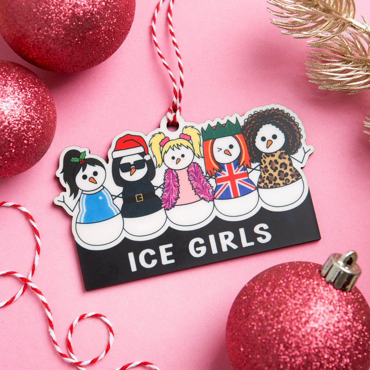 A rectangular Christmas ornament with rounded corners featuring cartoon snowmen dressed as The Spice Girls and the text 'ICE GIRLS' on a black background, hanging from a red-and-white striped string. The ornament is styled on a pink surface with glittery red ornaments, a gold pine branch, and candy canes.
