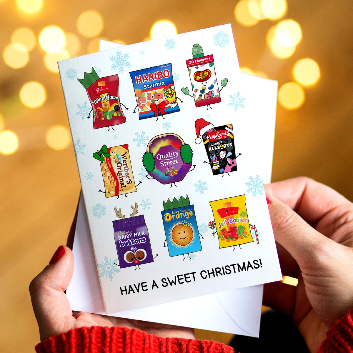 A person holds a white Christmas card featuring cartoon candy characters and the text 'HAVE A SWEET CHRISTMAS!'. The background is a warm, blurred bokeh of golden lights.
