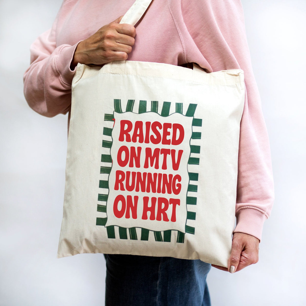 A person wearing a pink sweatshirt carries a cream canvas tote bag with a red and green graphic that reads 'RAISED ON MTV RUNNING ON HRT'.