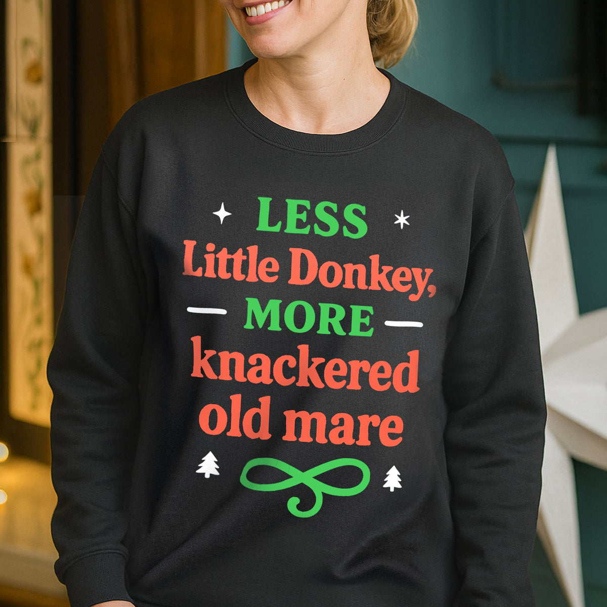 Person wearing a black Christmas sweatshirt with colourful text that says 'less little donkey, more knackered old mare' in a home setting 