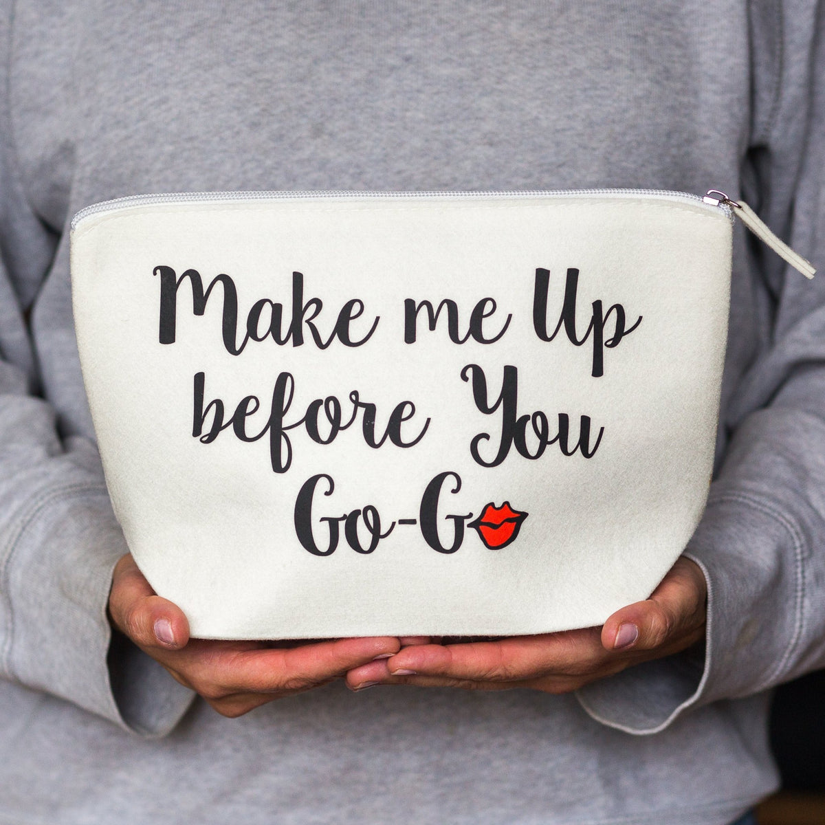 a cosmetic bag inspired the 80's wham hit and says 'Make me up befor you Go-Go' held in female hands against a grey jumper