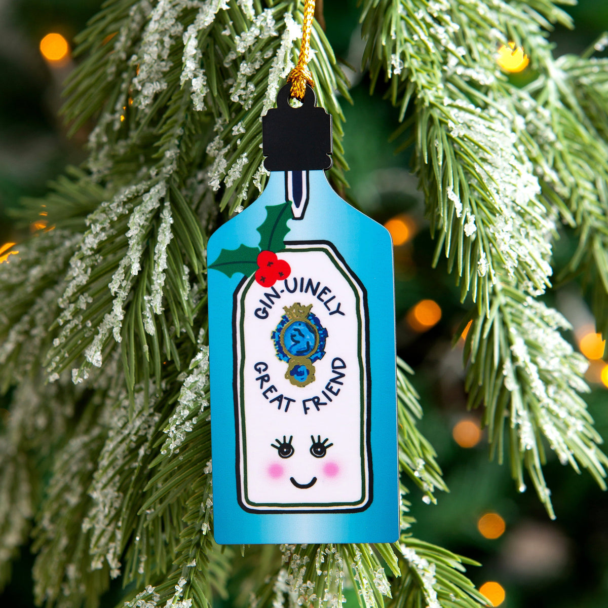 A Christmas tree ornament shaped like a cocktail glass, hanging from a gold string on a decorated Christmas tree. The ornament is light blue with a white label that reads 'ONLINELY GREAT FRIEND' with a cute face, and small holly accents.