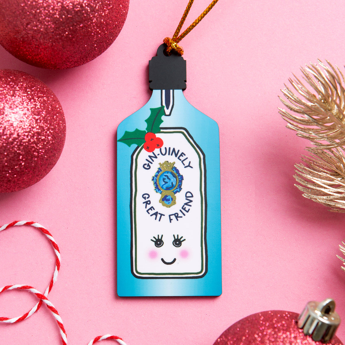 A Christmas ornament shaped like a Gin Tonic bottle, decorated with a blue gradient body, a white label reading 'GIN UINELY GREAT FRIEND,' a black cap, green holly leaves, red berries, and a cute face. It hangs from a gold string on a pink background with red glitter ornaments and gold pine branches around it.