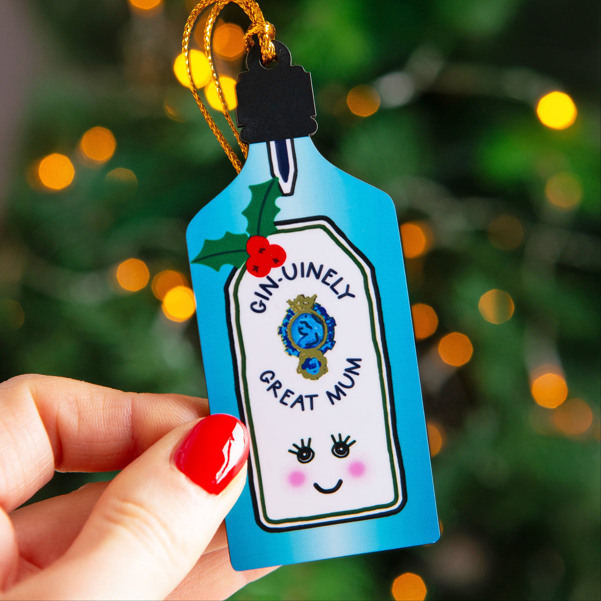 A hand with red nail polish holds a blue Christmas ornament shaped like a gin bottle. The ornament features a white label with the text 'GIN-UNELY GREAT MUM', a blue medallion, holly leaves, red berries, and a smiling face, with a gold hanging loop against a blurred green Christmas tree background with warm lights.