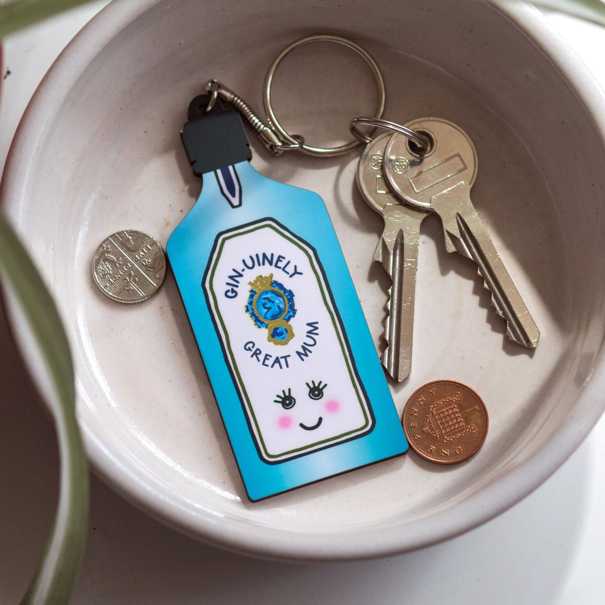 Keychain with a bottle design and text, placed on a ceramic dish with keys and coins.