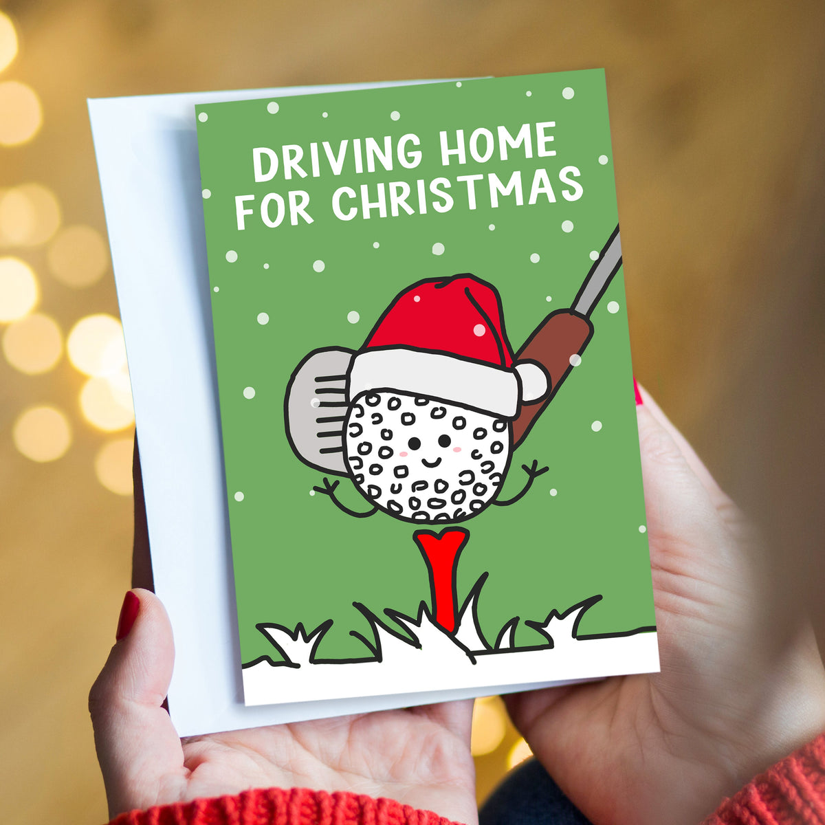 A person holds a Christmas greeting card with a white envelope behind it. The card has a green background with the text 'DRIVING HOME FOR CHRISTMAS' and an illustration of a golf ball wearing a Santa hat and using a golf club like a rake, with a red tee under its feet.