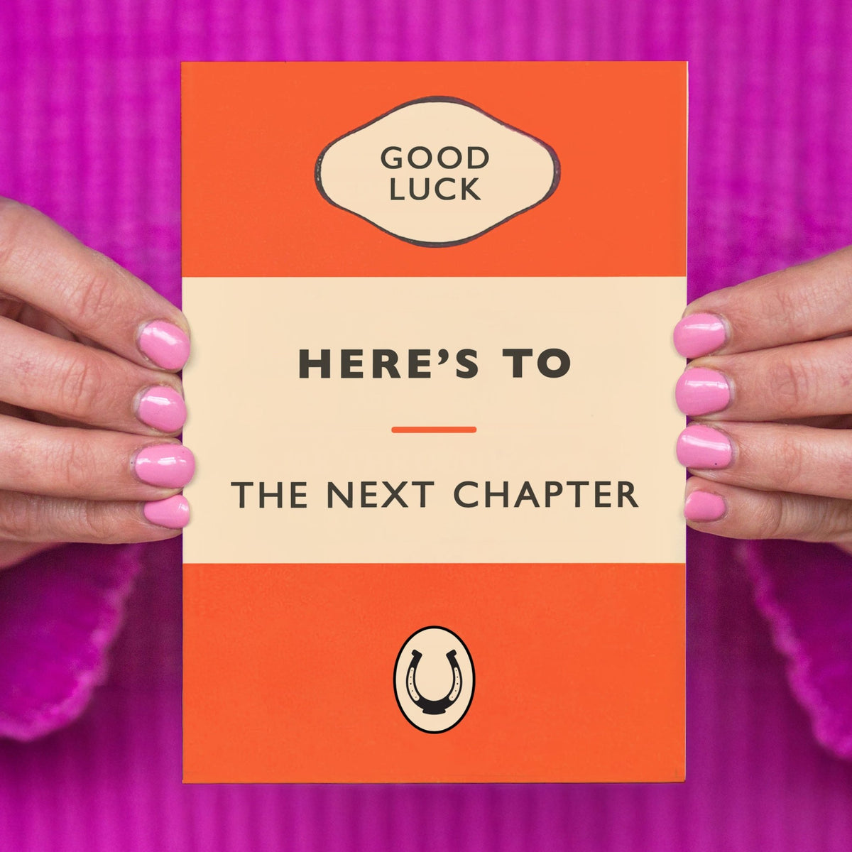 card designed to look like a book front cover that says good luck here's to the next chapter held in hands against a purple background