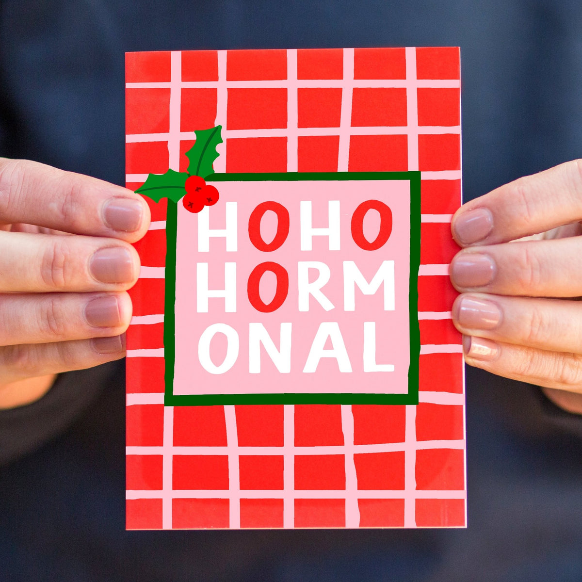 A person holds a square greeting card with a red and white grid pattern, featuring a central pink panel with the words 'HOHO HORMONAL' in red and white, plus a small green holly decoration in the top left corner.