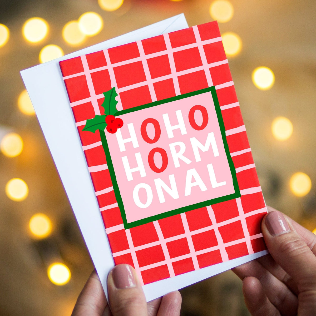 A person holds a red and white plaid greeting card with the words 'HOHO HORMONAL' in white letters on a pink center panel, decorated with a small green holly sprig. The background is softly blurred with warm string lights.