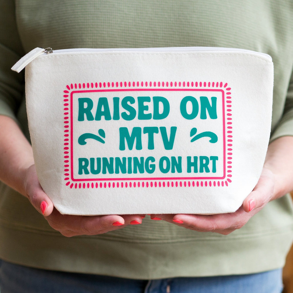 A person holding a white canvas zippered pouch with a pink and teal graphic that reads 'RAISED ON MTV RUNNING ON HRT'.