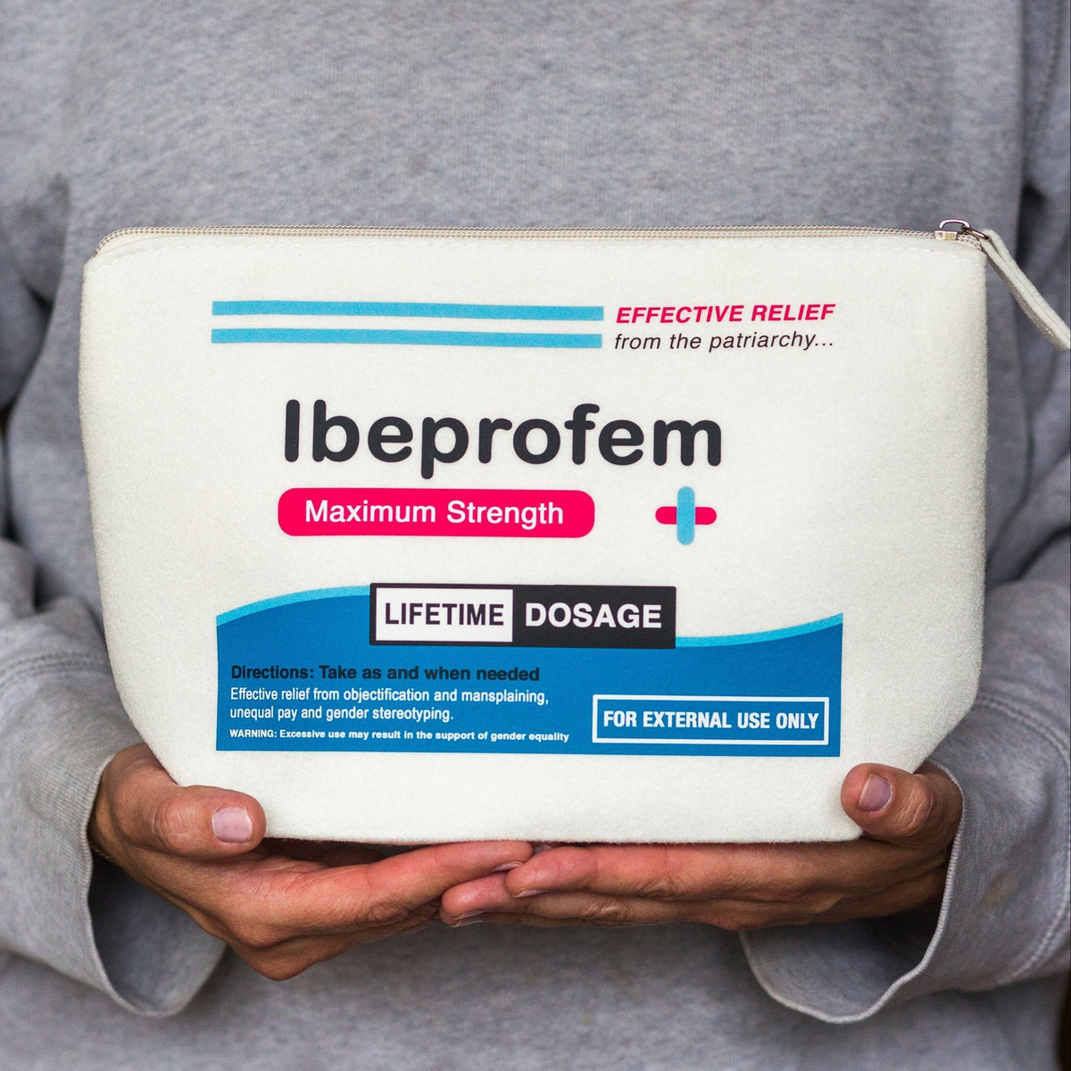 A person in a gray hoodie holds a white zippered pouch labeled 'Ibuprofen Maximum Strength' with blue and red text and graphics. The pouch is shown in a close-up shot against the person's torso.