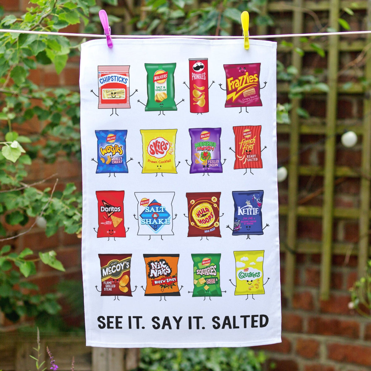 A white tea towel printed with colorful cartoon chip bag illustrations and the text 'SEE IT. SAY IT. SALTED'