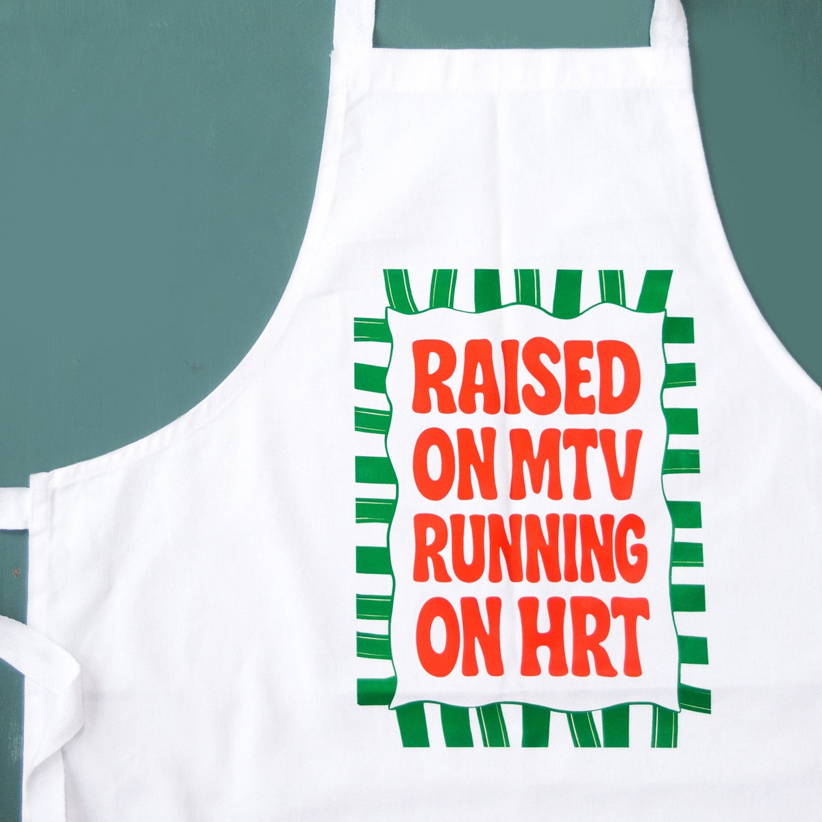 A white apron with a green and red graphic that reads 'RAISED ON MTV RUNNING ON HRT' on a teal background.