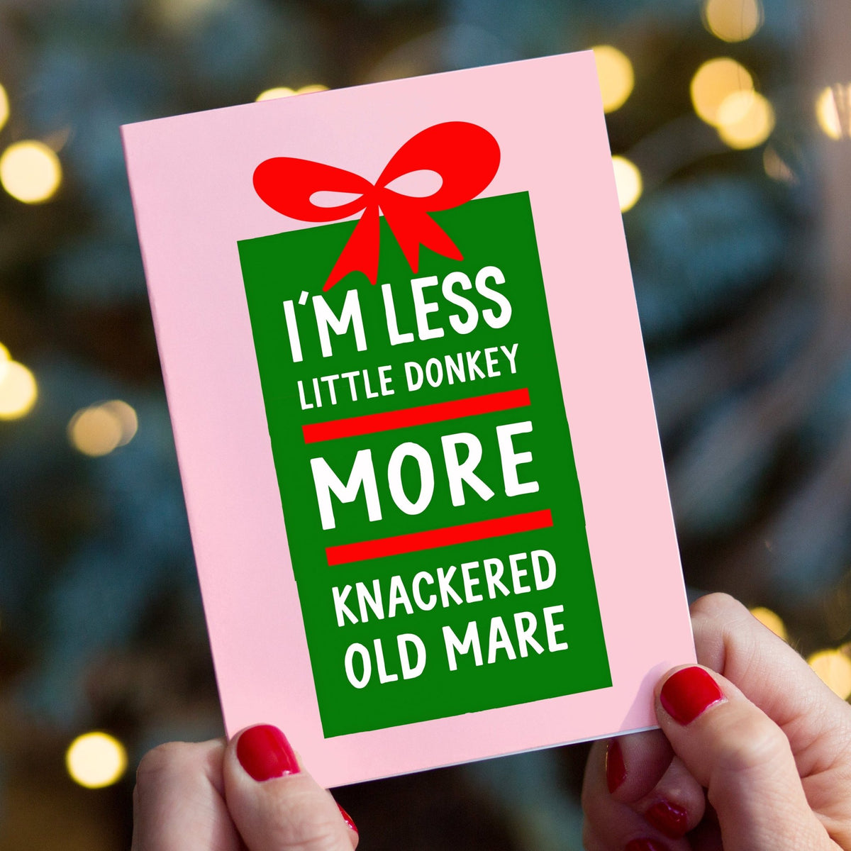 A person holds a pink greeting card with a green gift box graphic and red ribbon. The card reads, 'I'M LESS LITTLE DONKEY MORE KNACKERED OLD MARE' in white text.