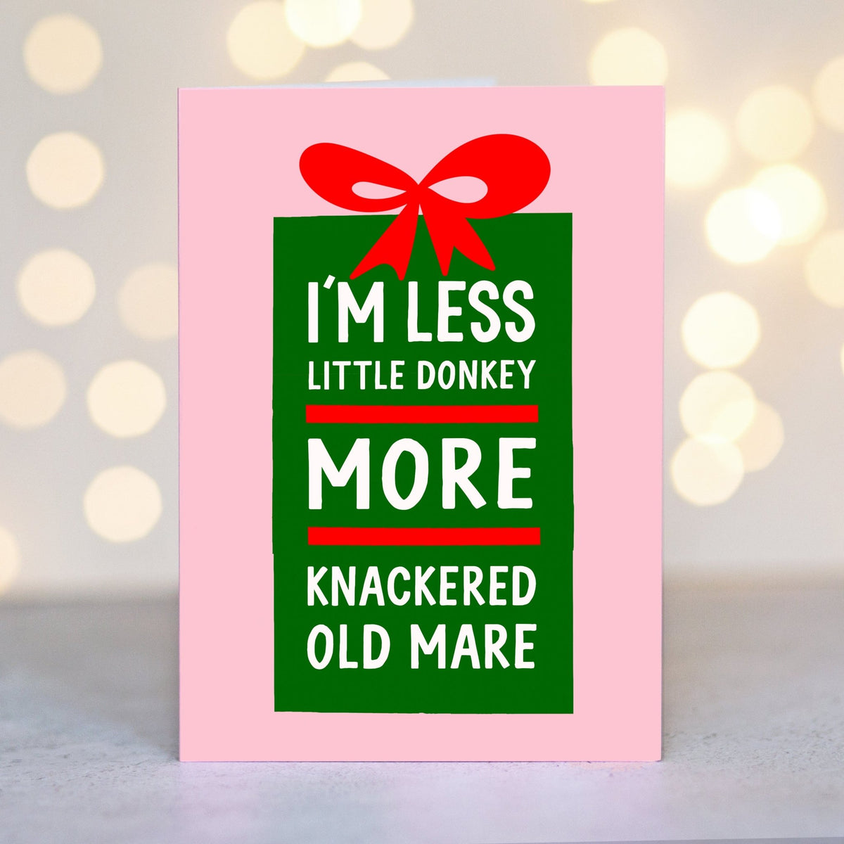A pink greeting card with a green gift box graphic and red ribbon, featuring the humorous text 'I'M LESS LITTLE DONKEY MORE KNACKERED OLD MARE' in white lettering, set against a soft blurred bokeh background.