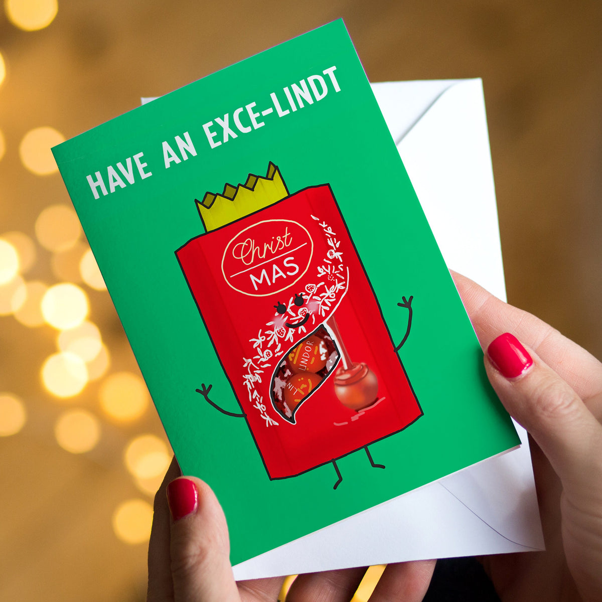 A person holds a green greeting card with a red cartoon box of Lindt chocolates illustration and the text 'HAVE AN EXCE-LINDT Christmas' above a white envelope. The background is softly blurred with warm bokeh lights.