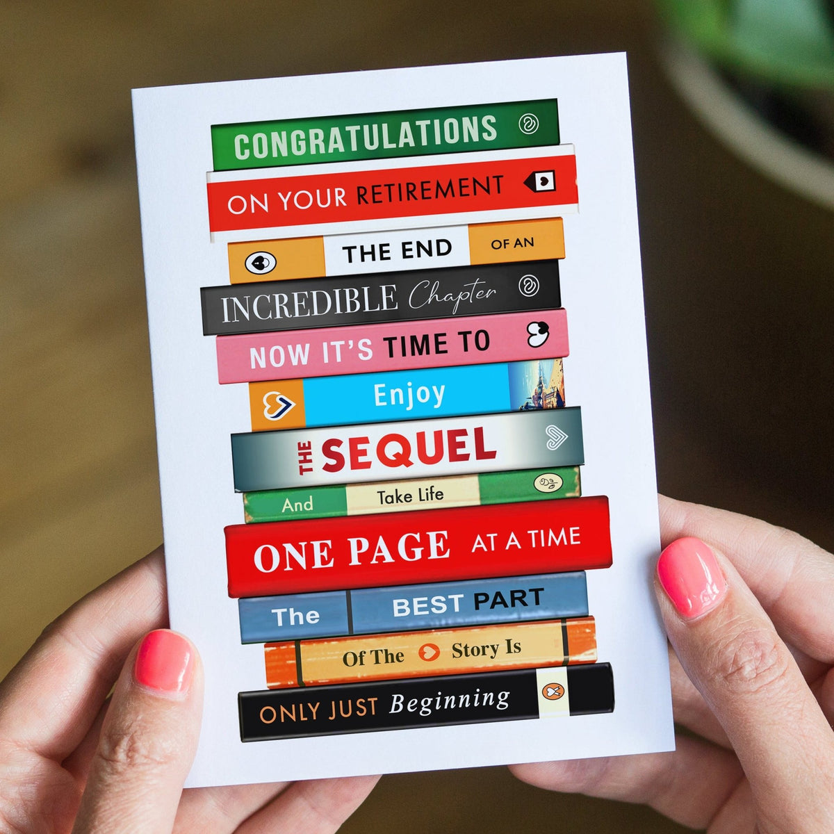 A person holds a white card printed with stacked colorful book spines and retirement-themed phrases like 'Congratulations on your retirement' and 'One page at a time.' The background is a softly blurred indoor setting with a wooden surface and greenery.