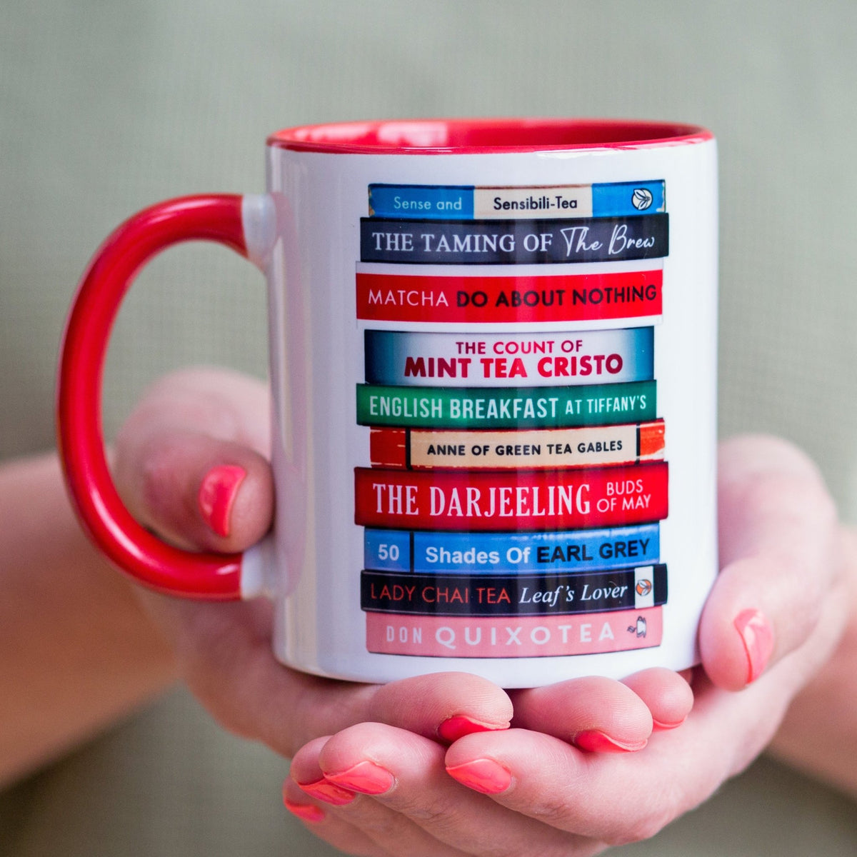 a white mug with a red handle that features a stack of books design that are each titled with a tea themedpun on literary book titles, held in female hands