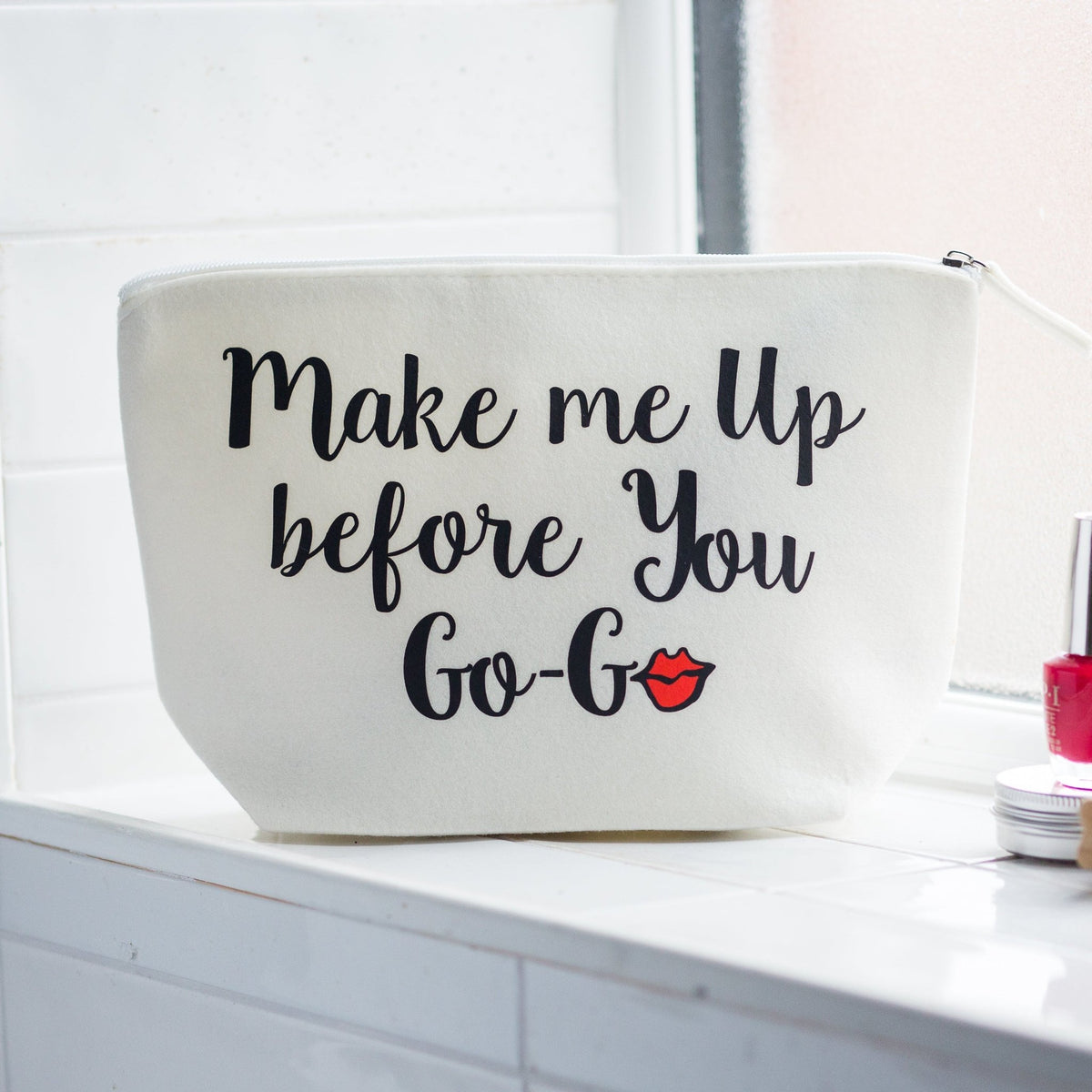 An off-white canvas cosmetic bag with the phrase 'Make me Up before You Go-G' in black script and a small red lips graphic, sitting on a white tiled counter next to beauty items.