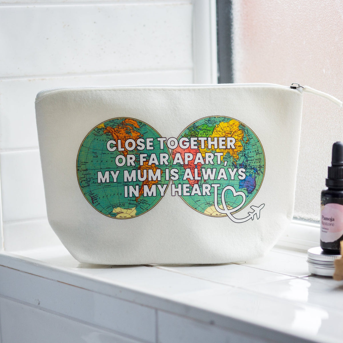 An off-white canvas cosmetic pouch with a zipper, featuring a graphic of two colorful world maps forming an infinity symbol with the text 'CLOSE TOGETHER OR FAR APART, MY MUM IS ALWAYS IN MY HEART'. The pouch is sitting on a white tiled windowsill with a small amber dropper bottle nearby.