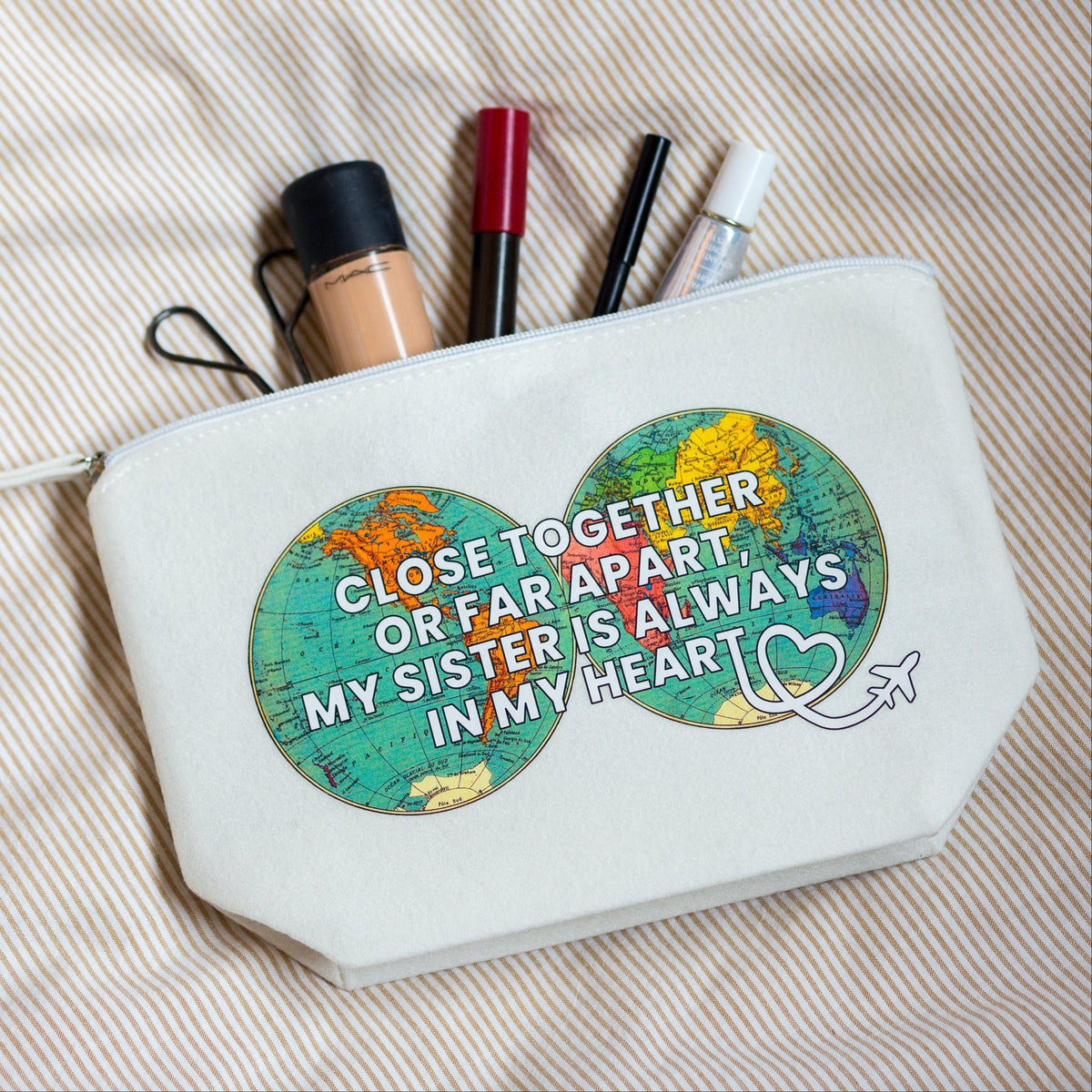 A white canvas cosmetic pouch with a zipper, featuring a graphic of two connected world maps with the text 'CLOSE TOGETHER OR FAR APART, MY SISTER IS ALWAYS IN MY HEART' in white. The pouch is shown filled with makeup items and lies on a beige and white striped fabric background.