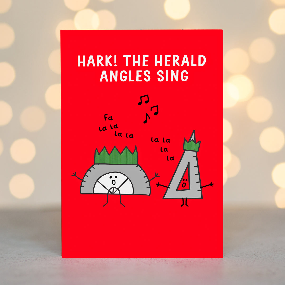 A red christmas card with a humorous illustration of a steering wheel and a speedometer character singing 'la la la', with the text 'HARK! THE HERALD ANGLES SING'. The card is photographed standing upright against a soft-focus background of warm bokeh lights.