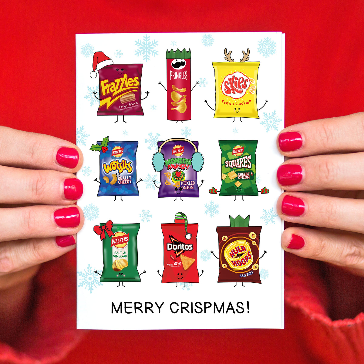 A person with bright pink fingernails holds a white Christmas card featuring cartoon illustrations of various branded crisp packets with arms and legs, with the text 'MERRY CRISPMS!'. The background is a solid bright red sweater.