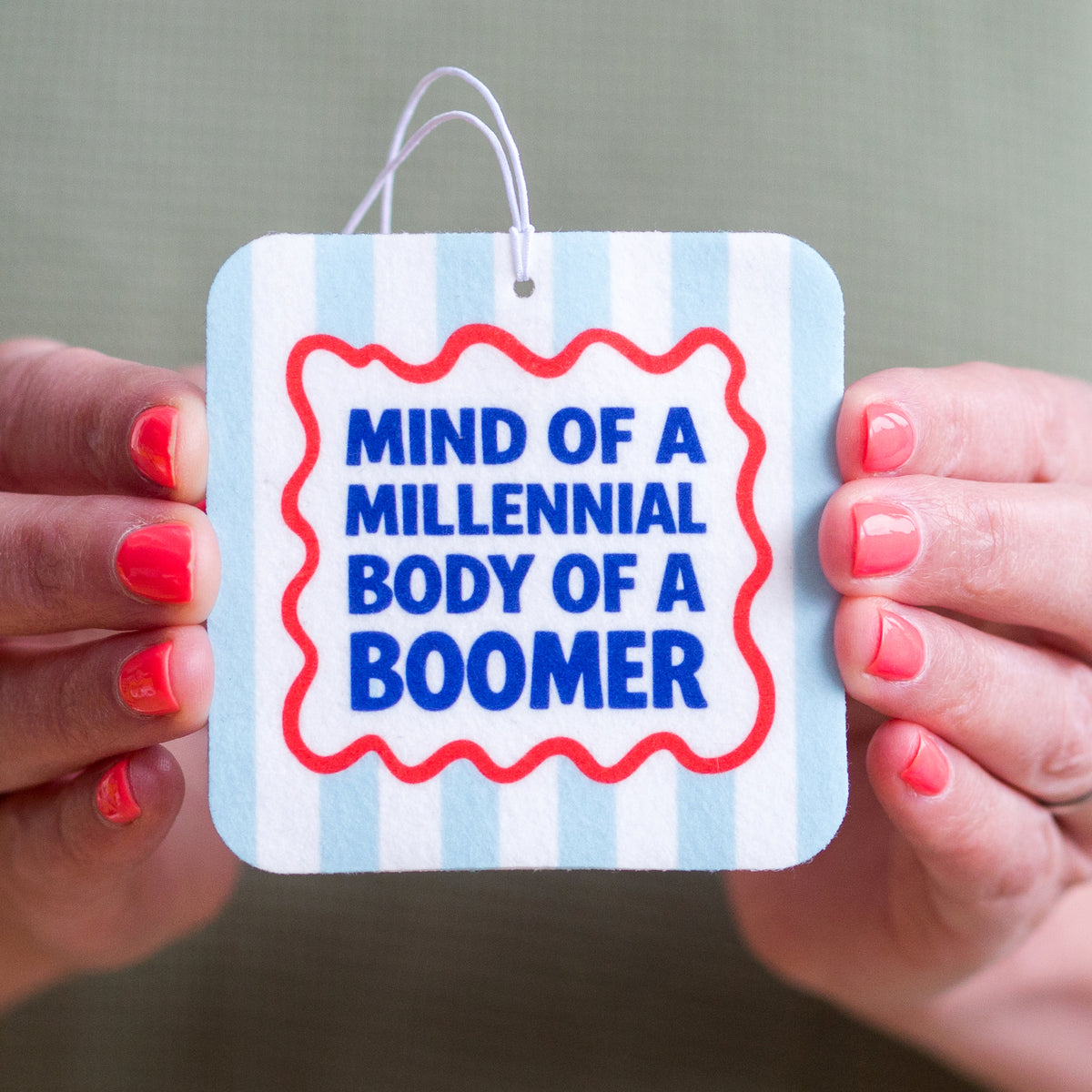 Hand holding a car air freshener with 'Mind of a Millennial, Body of a Boomer' text on a gray background