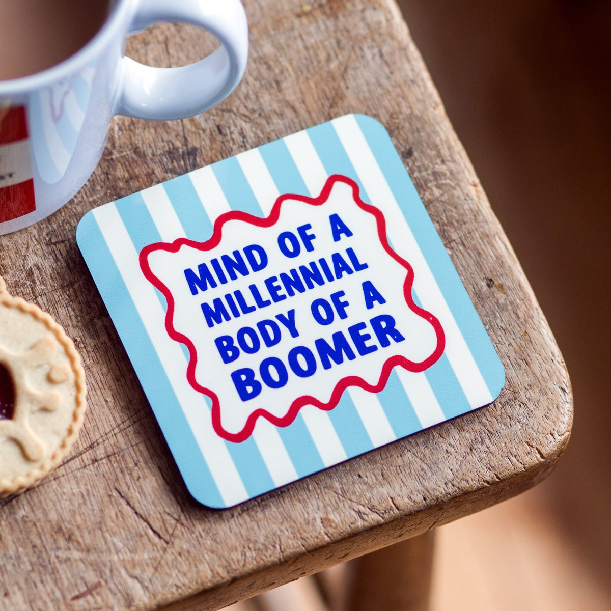 Coaster with text 'Mind of a Millennial Body of a Boomer' on a wooden surface with a cup and cookie.