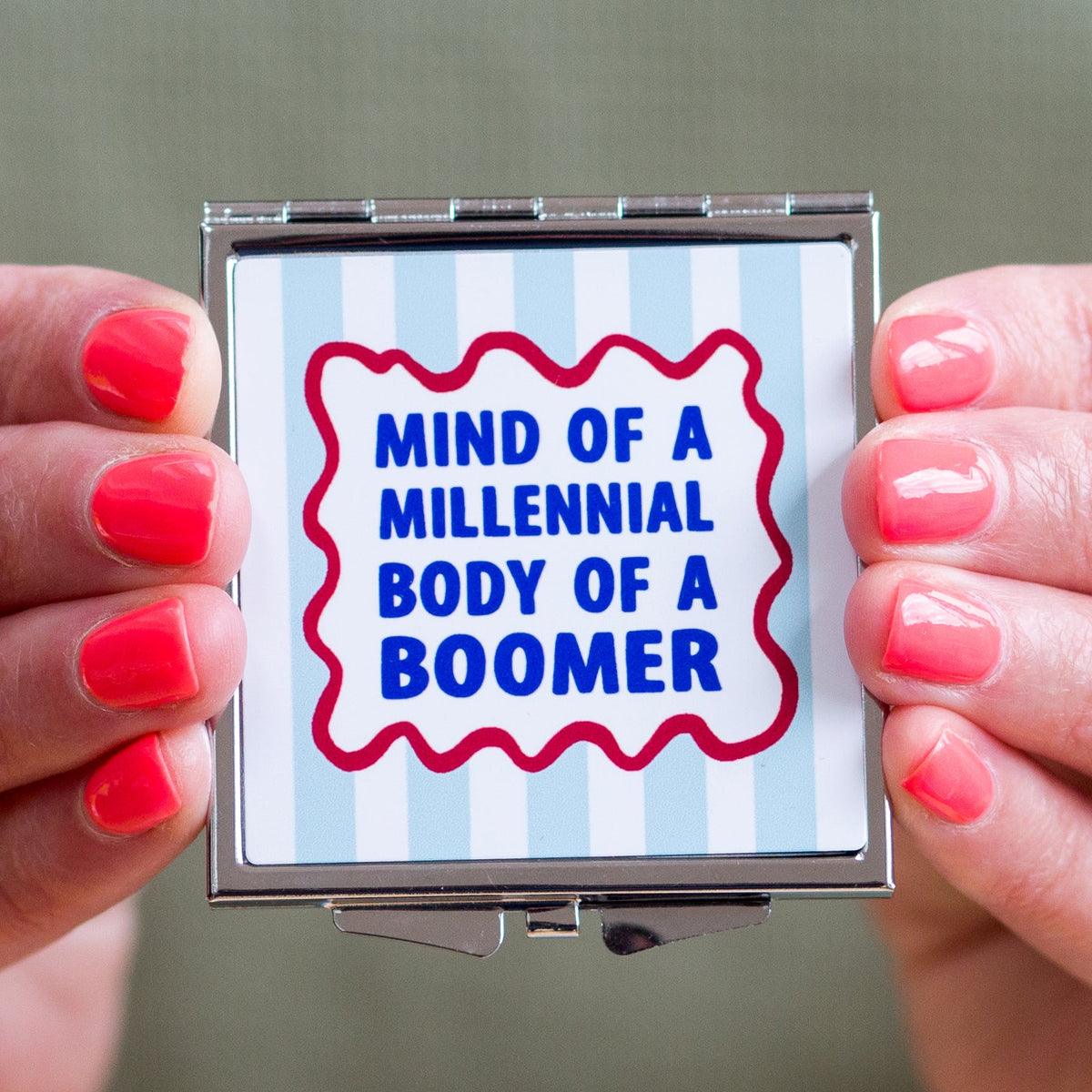 Compact mirror with 'Mind of a Millennial, Body of a Boomer' text held by hands with red nail polish.