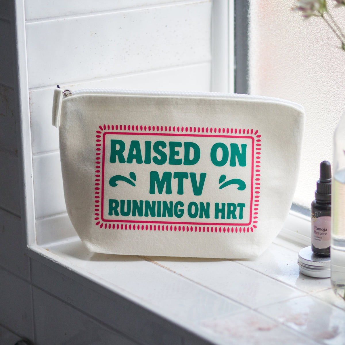 An off-white canvas cosmetic pouch with a green and red graphic reading 'RAISED ON MTV RUNNING ON HRT' sits on a tiled bathroom windowsill next to a small dropper bottle.