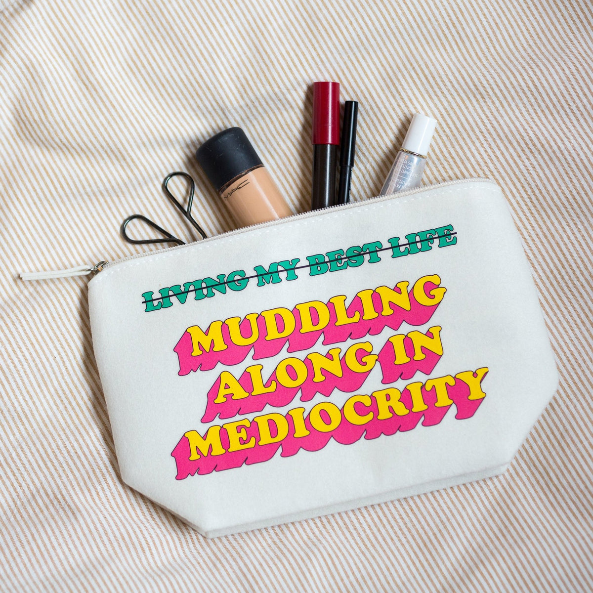 A white canvas cosmetic pouch with colorful retro text reading 'LIVING MY BEST LIFE MUDDING ALONG IN MEDIOCRITY' lies on a beige and white striped fabric background, with makeup items peeking out from the top.