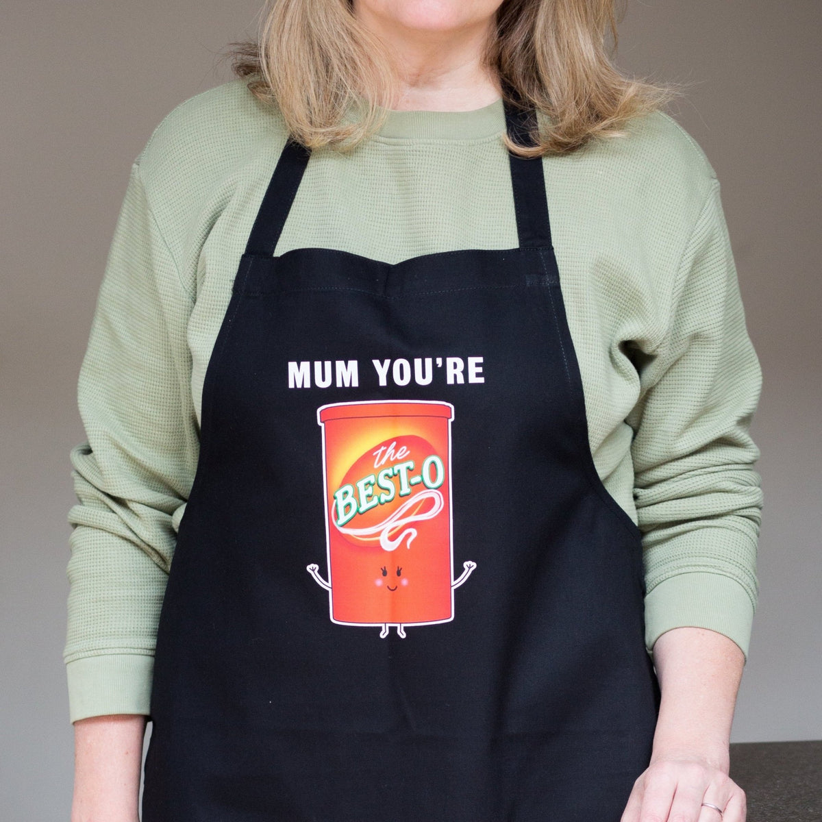 black apron that says mum you're the best-o on an illustration of a jar of Bisto, being worn by a woman in a kitchen