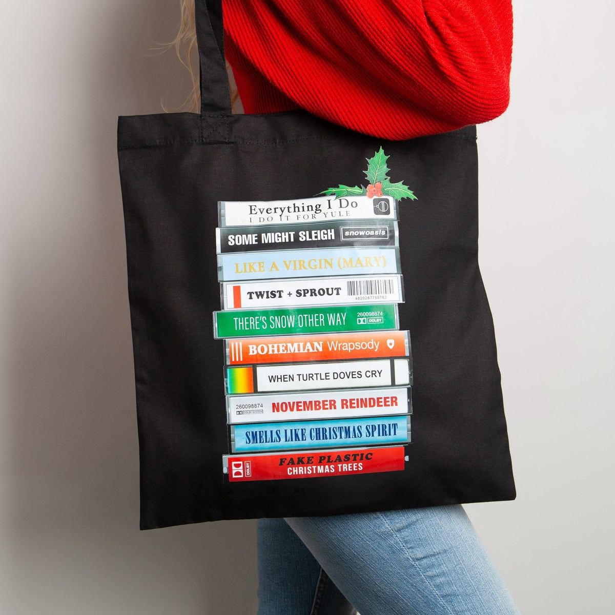 A woman wearing a red sweater and blue jeans carries a black canvas tote bag with a colorful graphic of stacked cassette tapes with festive puns on classic song titles, set against a plain light gray wall.