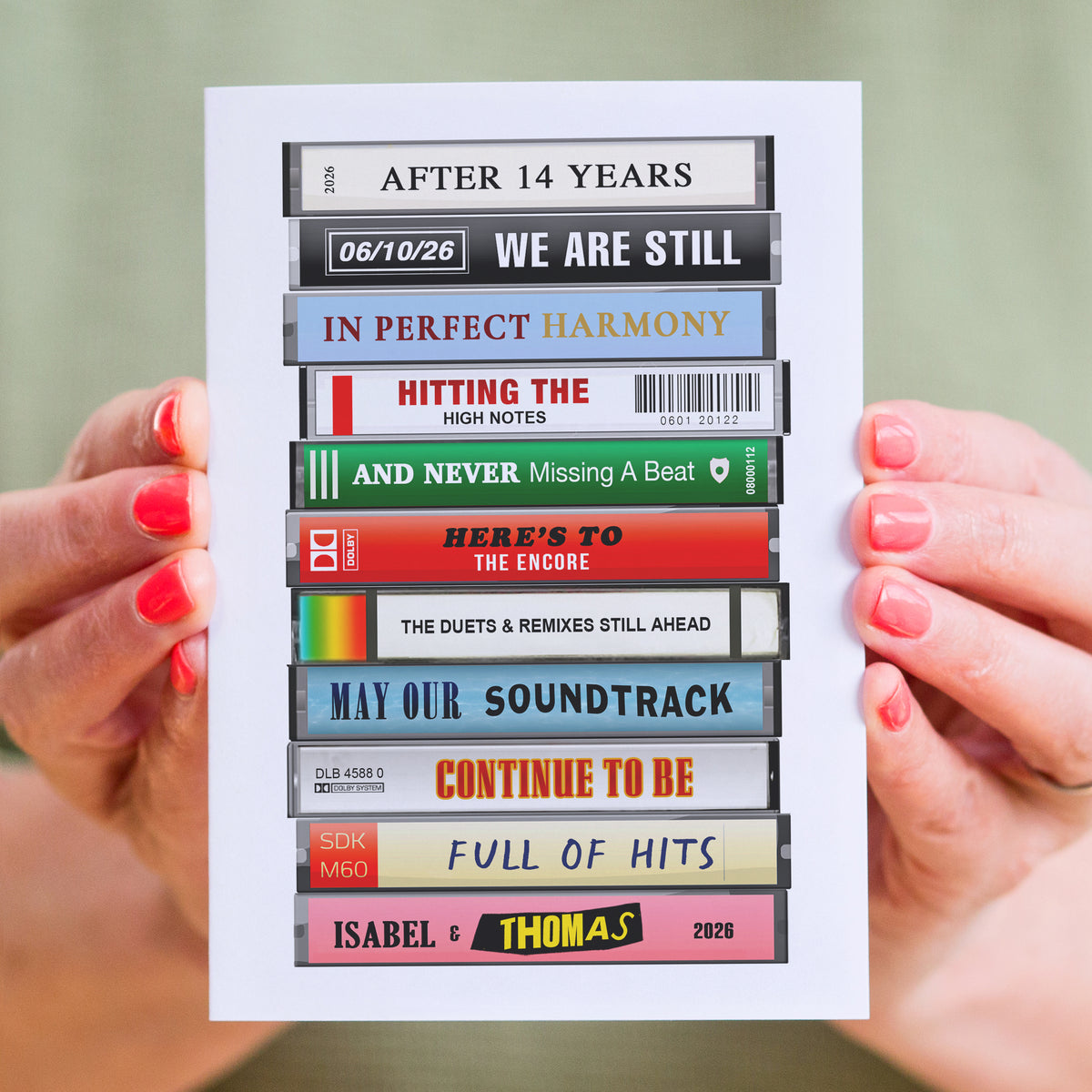 A white rectangular card held by two hands with pink nail polish. The card features a graphic of stacked cassette tapes with various song titles and phrases printed on them, such as 'AFTER 14 YEARS', 'WE ARE STILL', 'IN PERFECT HARMONY', 'HITTING THE HIGH NOTES', 'AND NEVER Missing A Beat', 'HERE'S TO THE ENCORE', 'MAY OUR SOUNDTRACK', 'CONTINUE TO BE', 'FULL OF HITS', and 'ISABEL & THOMAS 2028'.