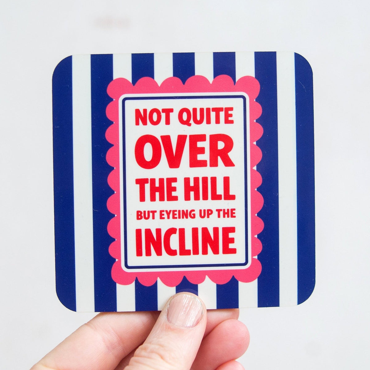 Coaster with humorous text 'Not quite over the hill but eyeing up the incline' held by a hand against a white background.