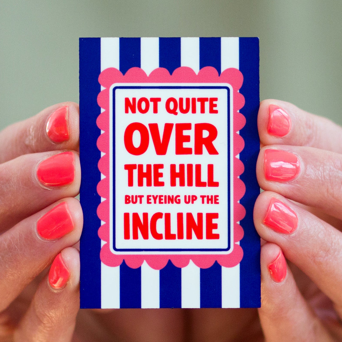 Fridge magnet with text 'Not quite over the hill but eyeing up the incline' held by hands with red nail polish.
